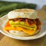 A toasted English muffin breakfast sandwich with melted cheddar cheese, a folded fried egg, and spicy sautéed kimchi on a small plate.
