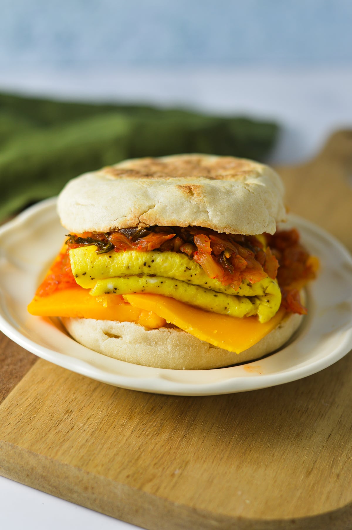 A toasted English muffin breakfast sandwich with melted cheddar cheese, a folded fried egg, and spicy sautéed kimchi on a small plate.