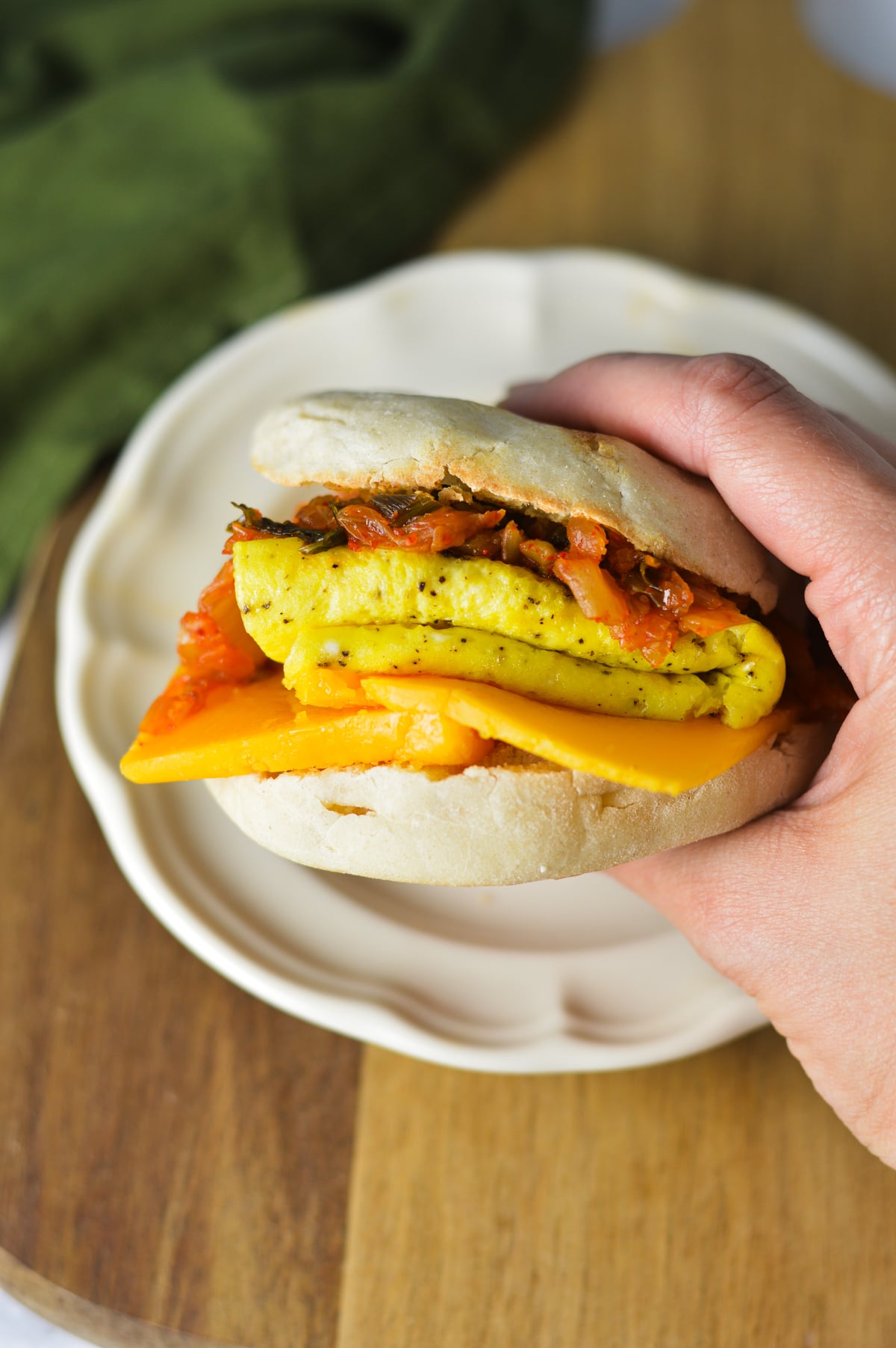 A hand holding a freshly assembled kimchi breakfast sandwich, showing the layers of fluffy cooked egg, melted cheddar cheese, and sautéed kimchi inside a toasted English muffin.