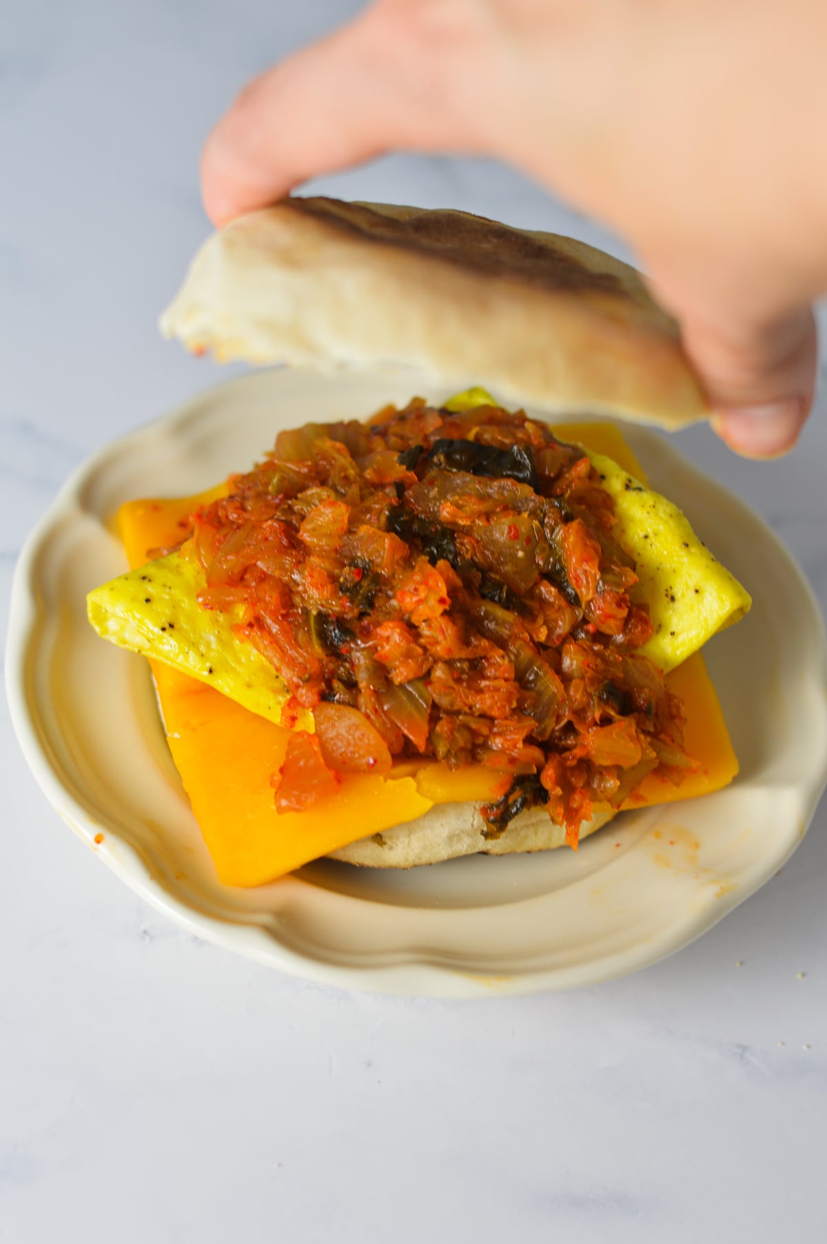 A hand placing a toasted slice of English muffin on a Kimchi Breakfast Sandwich.