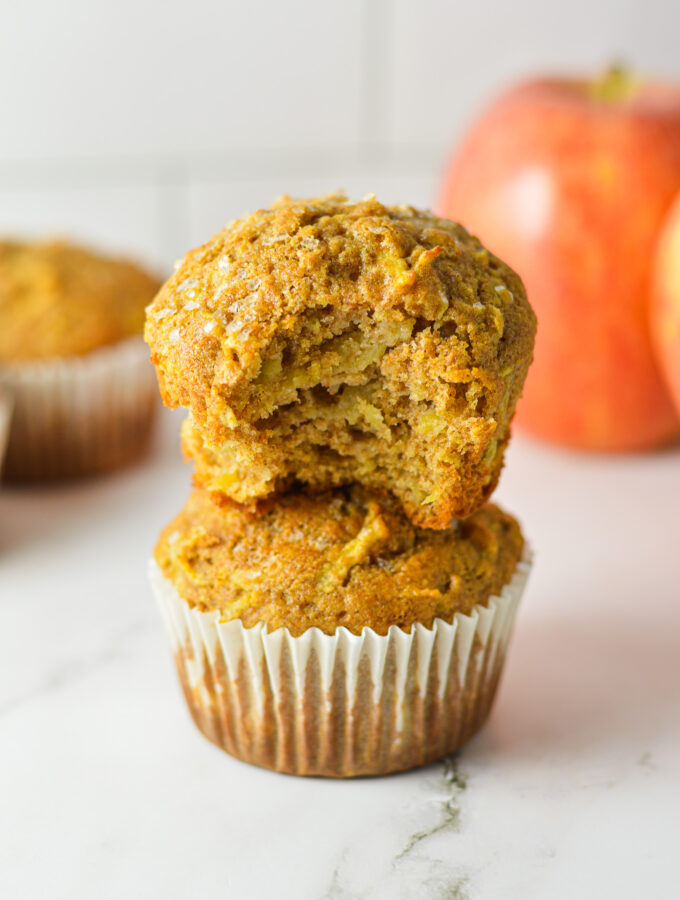 A stack of Whole Wheat Apple Muffins, with the paper liner taken off the top moist muffin and a bite taken out showing the soft inside.