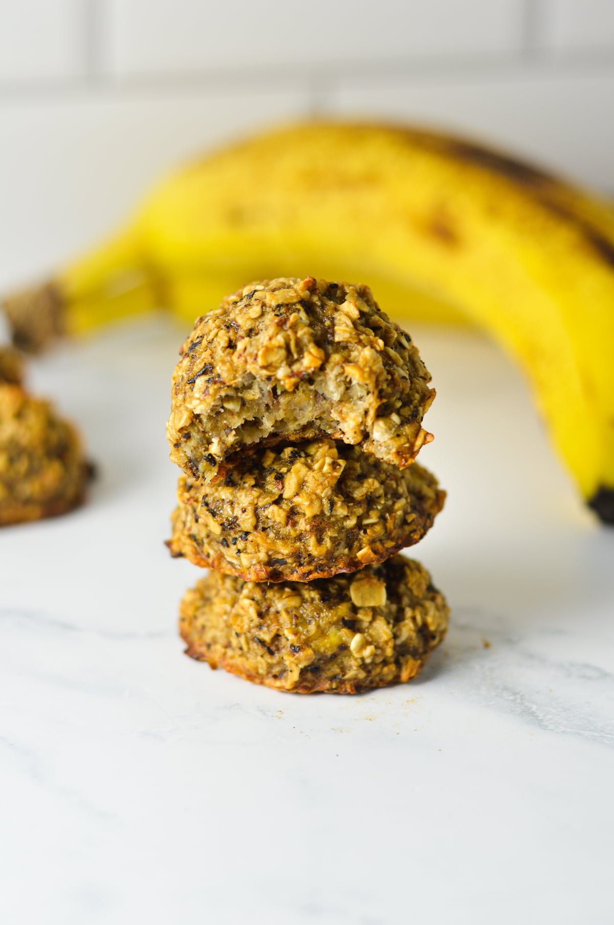 Stack of eggless banana chai latte oat cookies sitting next to a ripe yellow banana on a marble surface.