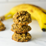 Three eggless banana chai latte oat cookies stacked in a vertical tower next to a ripe yellow banana on a marble countertop.