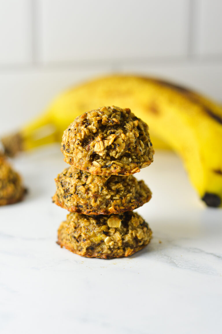 Three eggless banana chai latte oat cookies stacked in a vertical tower next to a ripe yellow banana on a marble countertop.