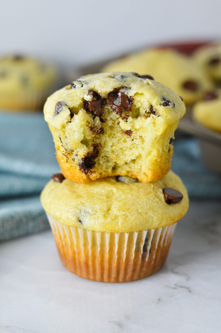 A stack of delicious Sour Cream Chocolate Chip Muffins with a bite taken out of the top muffin, showing its fluffy texture and melty chocolate chips.