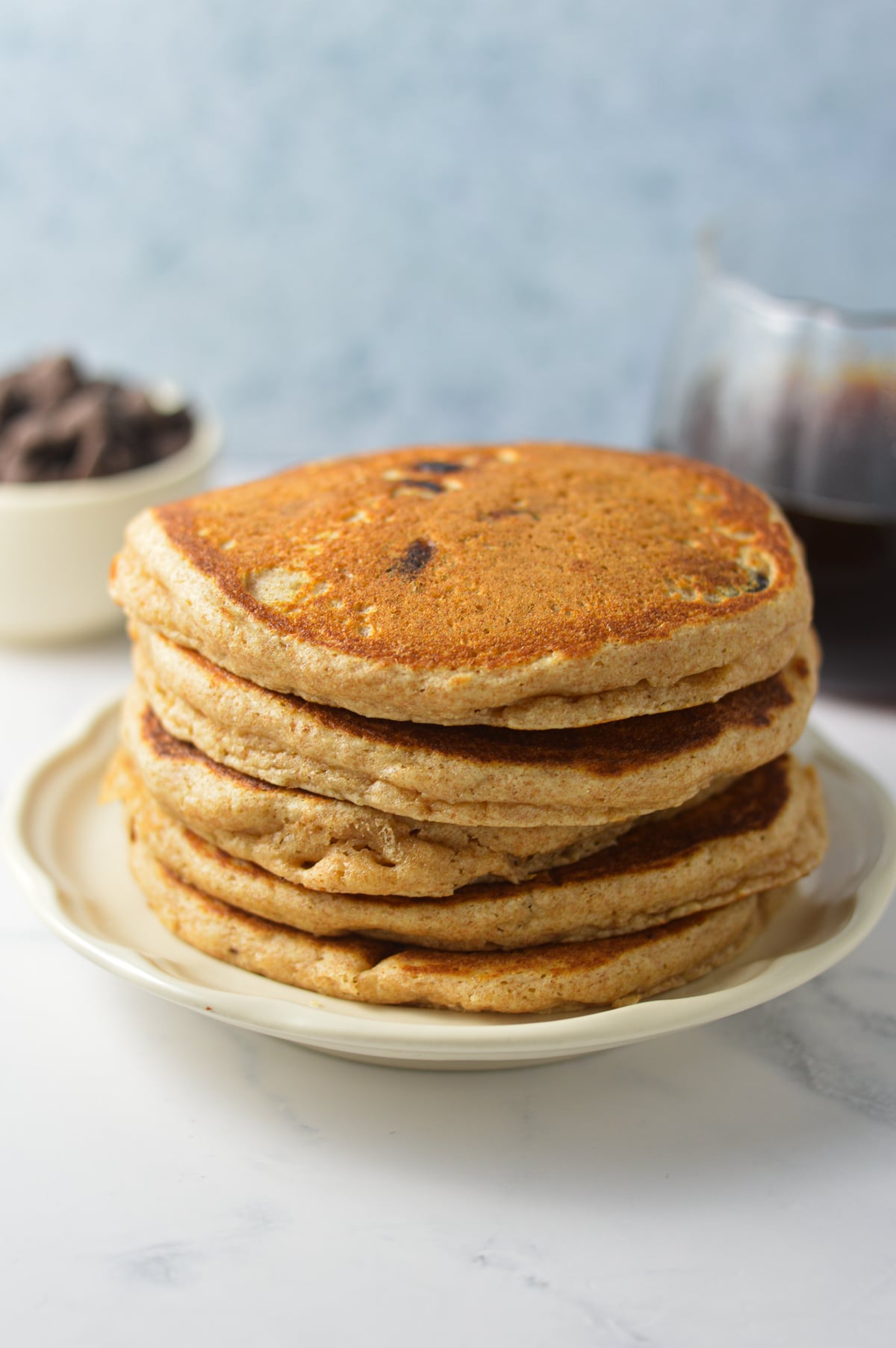 A stack of Whole Wheat Chocolate Chip Pancakes, with some semi-sweet chocolate chips and a cup of coffee in the background.