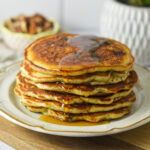 A stack of golden-brown chocolate chip banana pecan pancakes drizzled with maple syrup, presented on a white plate next to a small potted plant on a wooden surface.