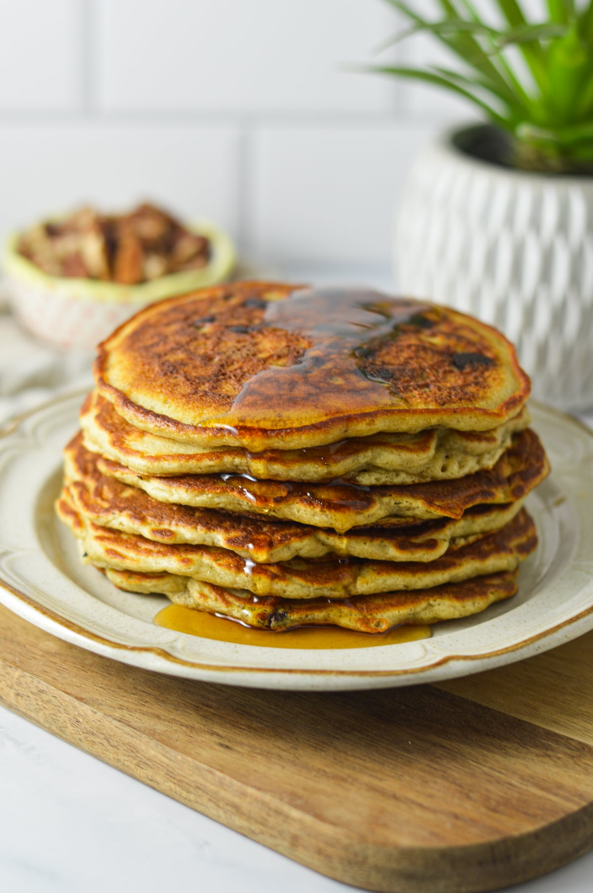 A stack of golden-brown chocolate chip banana pecan pancakes drizzled with maple syrup, presented on a white plate next to a small potted plant on a wooden surface.