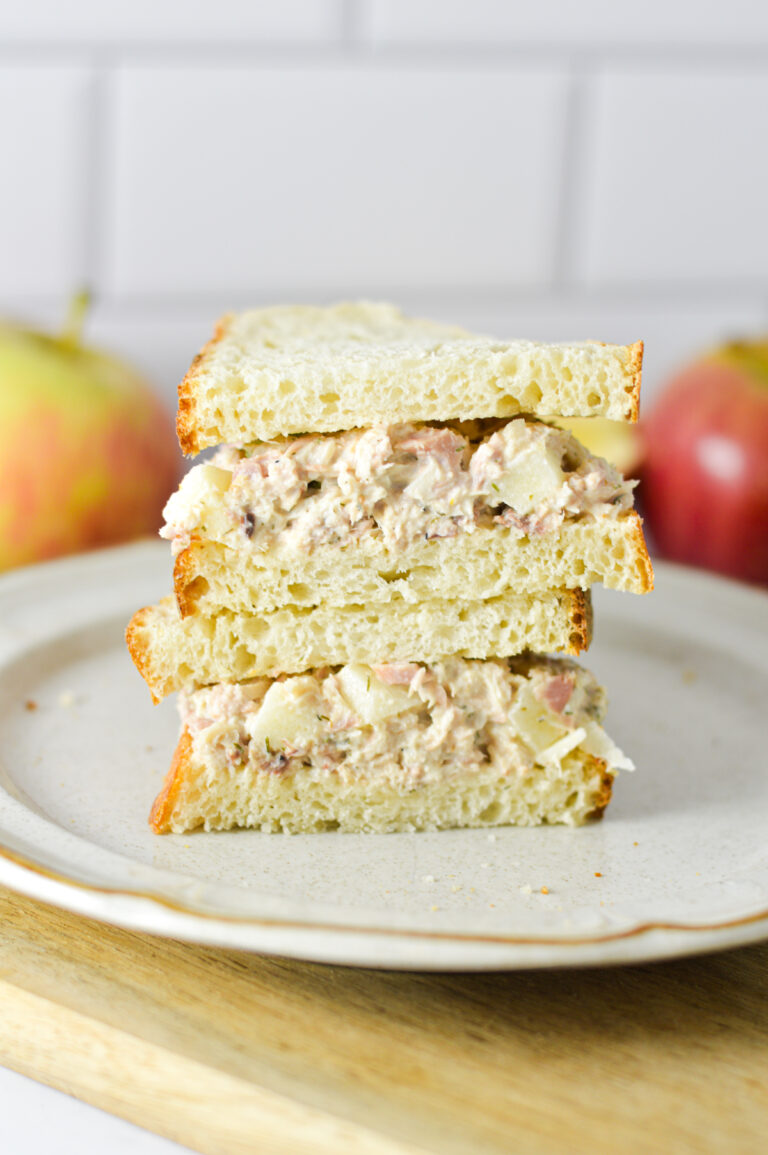 Apple Tuna Salad Sandwich sliced in half and stacked on a plate, with some fresh apples in the background.