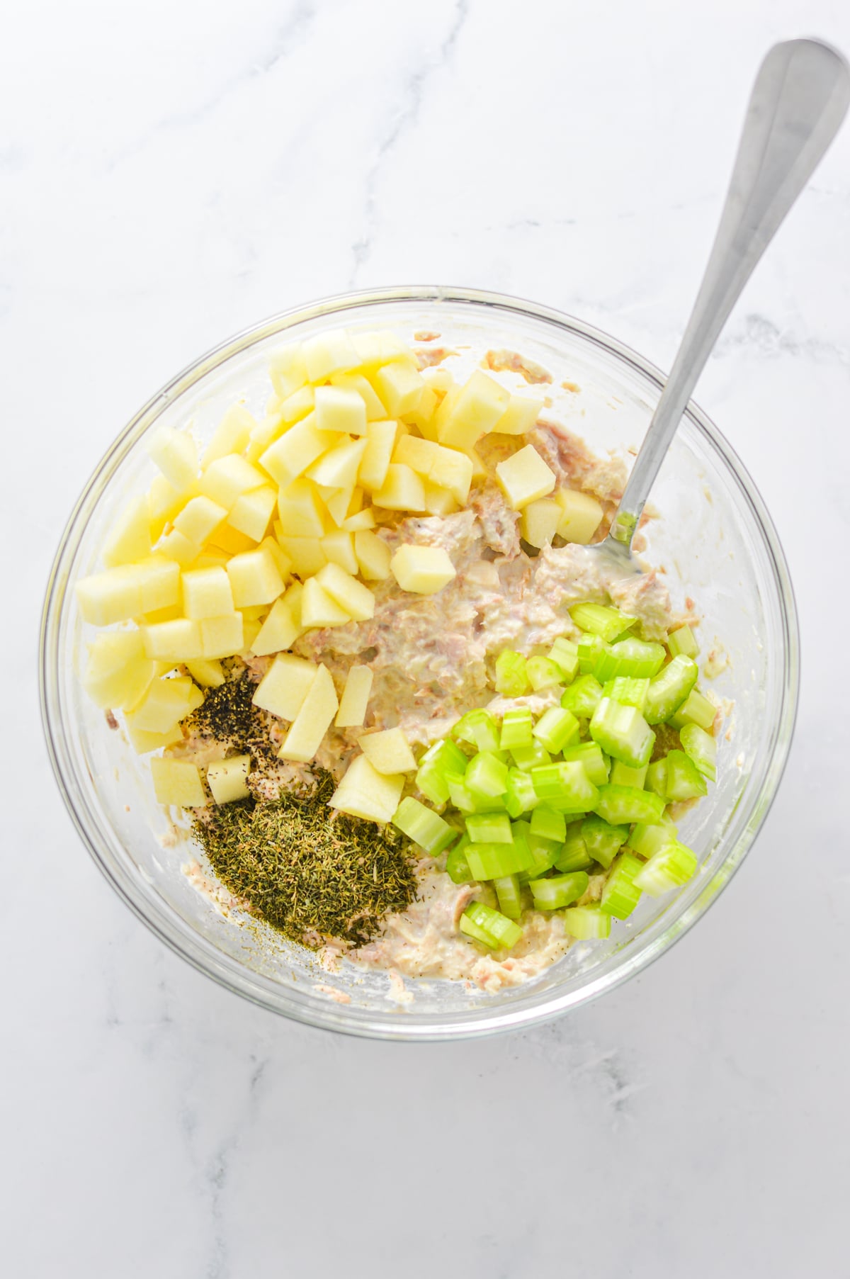A glass bowl with tuna, mayo, lemon juice, diced apples, celery, dried dill and salt and pepper about to be mixed with a metal spoon.