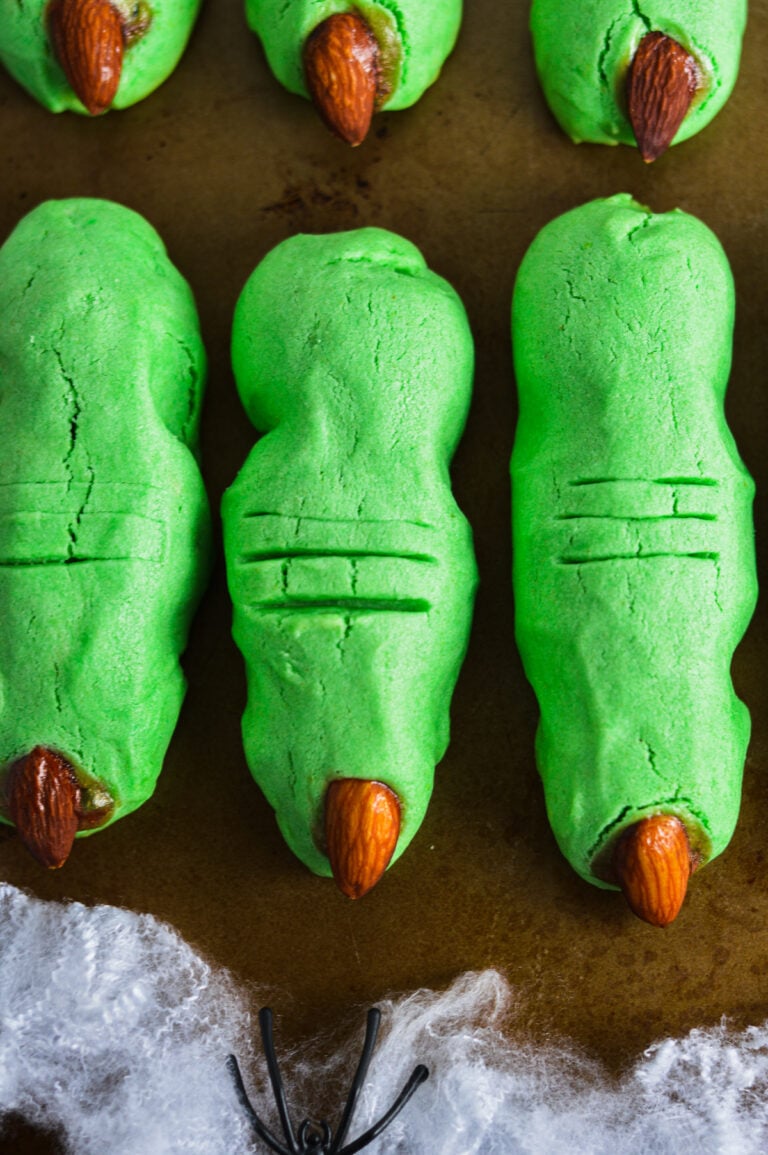 Green witch finger cookies with almond fingernails and red jam around the edges, arranged on a baking sheet.