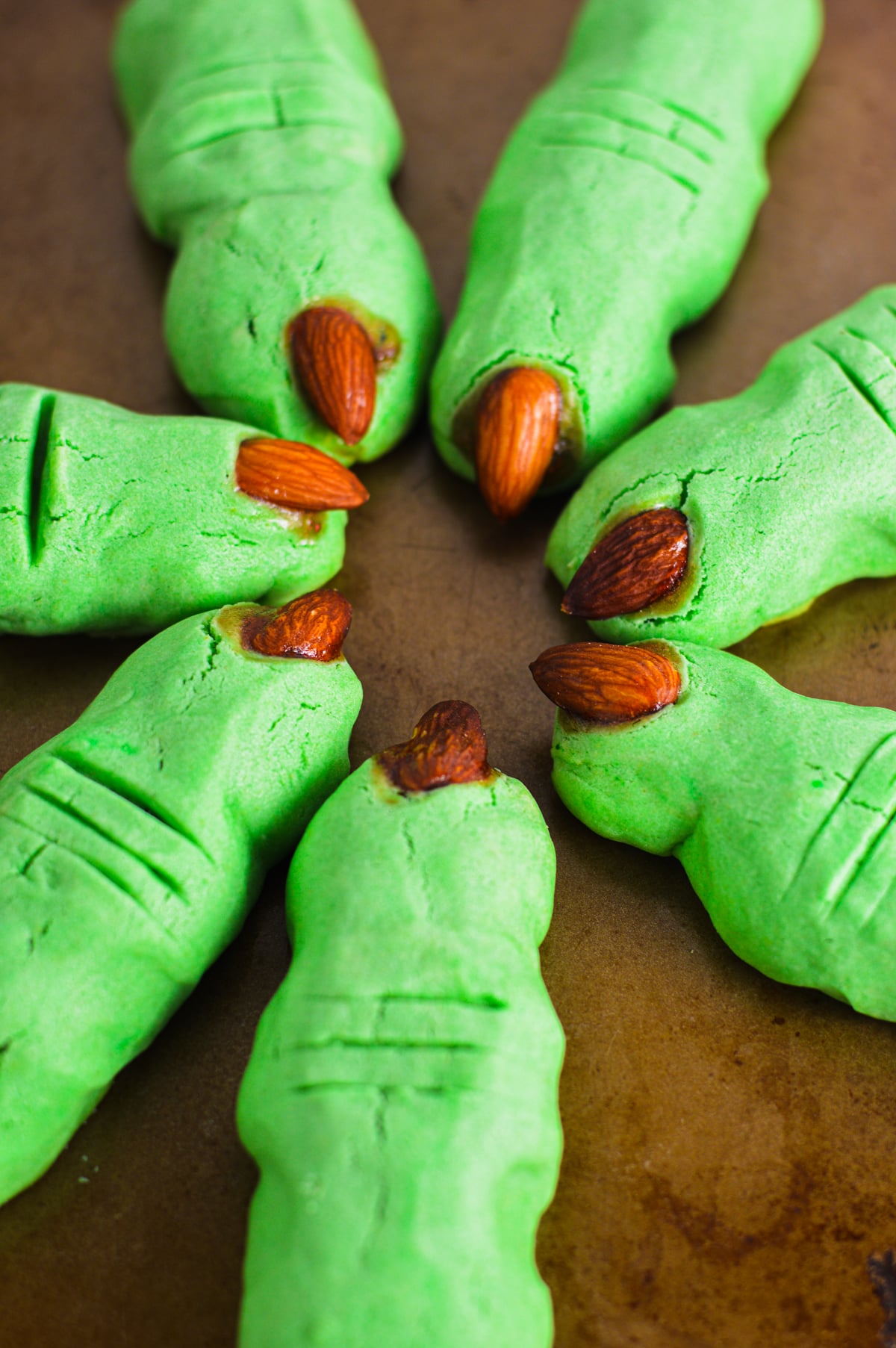 Spooky green witch finger cookies with almond fingernails and strawberry jam detail, arranged in a circular pattern on a cookie sheet.