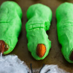 Spooky green witch finger cookies with almond fingernails and red jam accents, arranged next to a plastic spider on pretend spiderwebs.