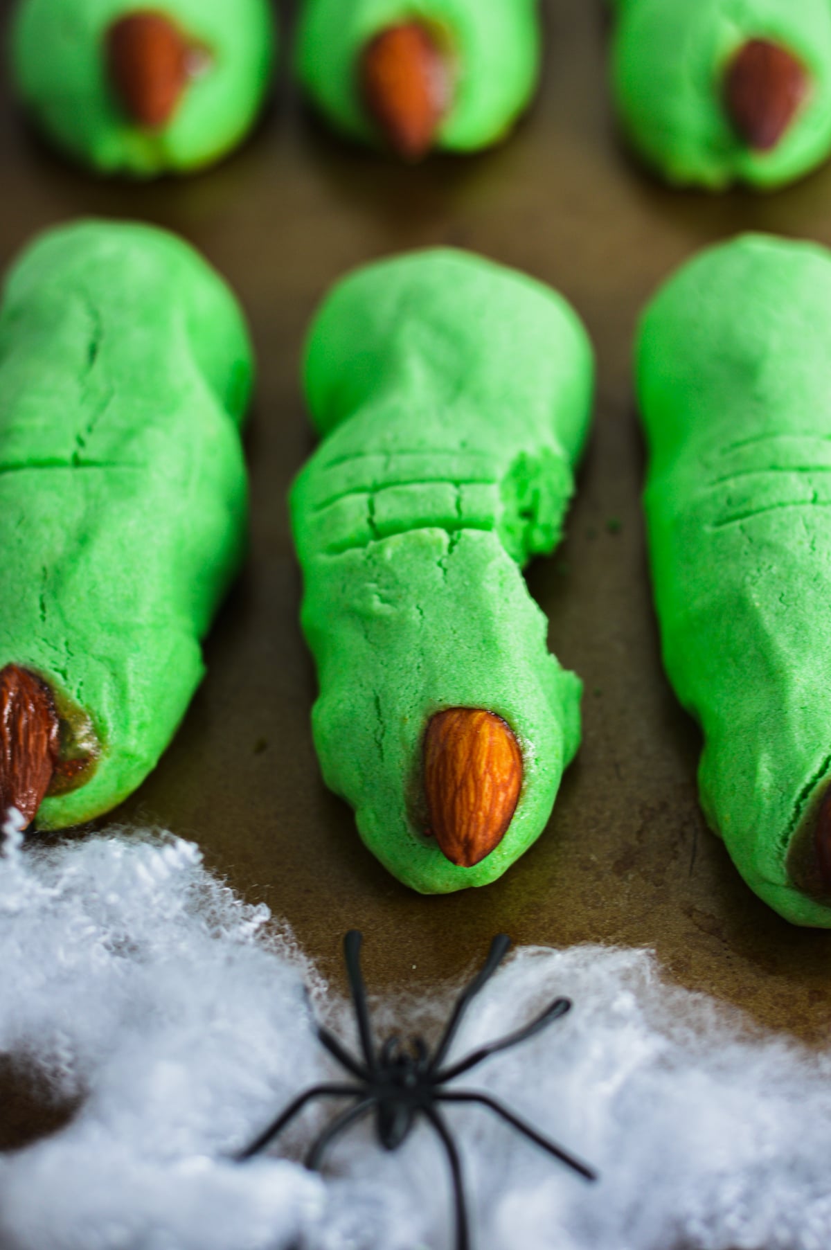 Spooky green witch finger cookies with almond fingernails and red jam accents, arranged next to a plastic spider on pretend spiderwebs.
