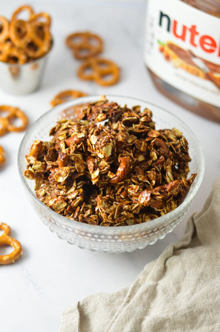 A small bowl filled with Pretzel and Nutella Granola topped with pretzel pieces, with a jar of Nutella and loose pretzels in the background.