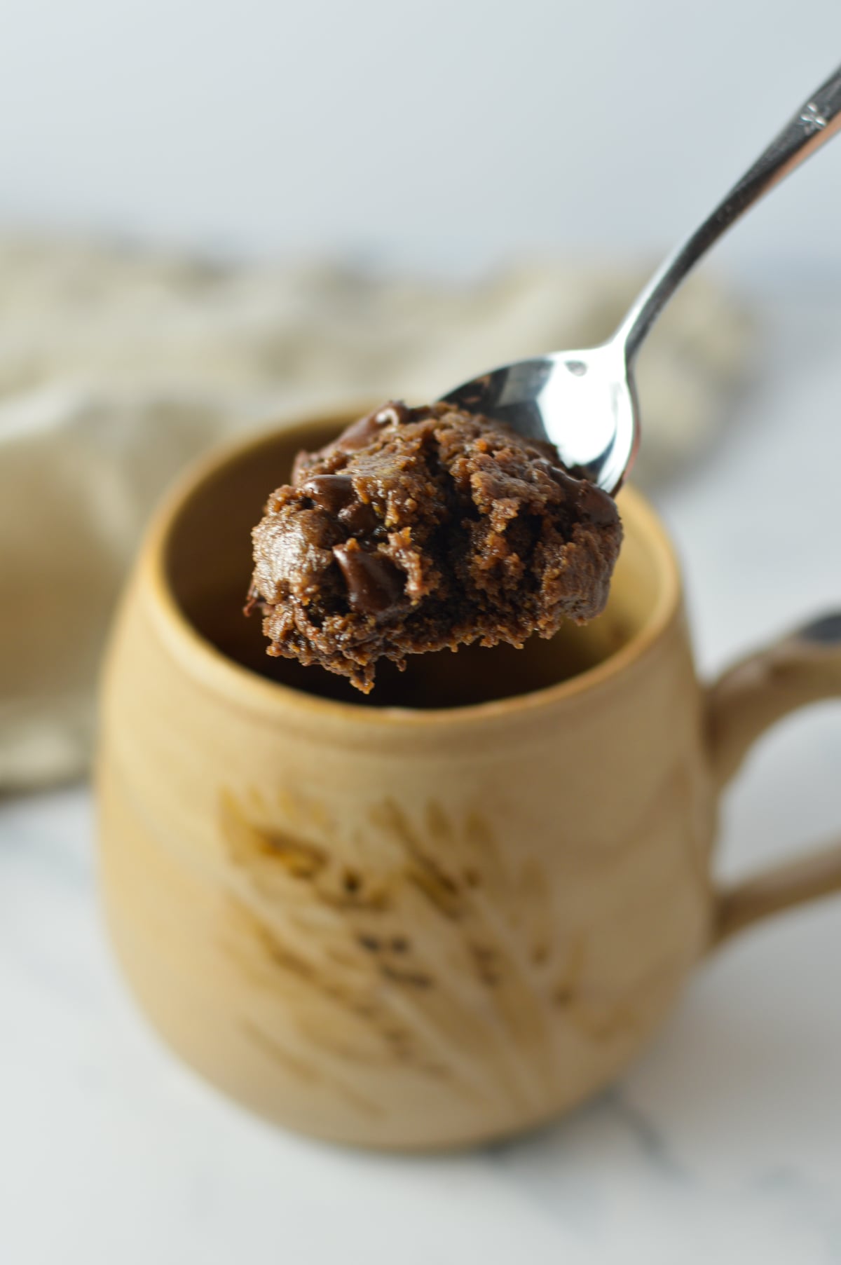 A close-up of a warm, fudgy microwave brownie in a tan ceramic mug with a spoonful being lifted out.