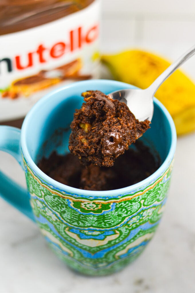 A spoon taking a bite of Banana Nutella Mug Cake in a ceramic mug, showing the moist texture with some chunks of banana in the chocolate cake.