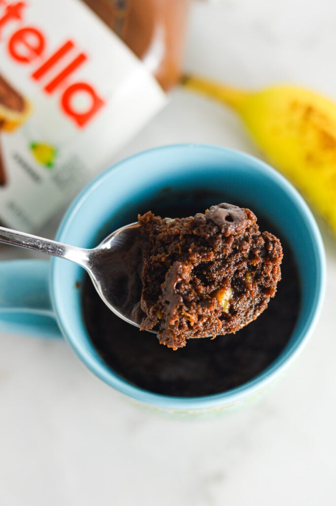 A spoon taking a bite of Banana Nutella Mug Cake from a large mug filled with the moist cake.