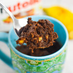 A small spoon taking a bite of Banana Nutella Mug Cake from a microwave safe mug, with the rich texture and small banana pieces showing in the chocolate cake.