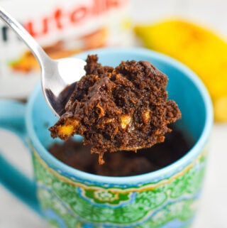 A small spoon taking a bite of Banana Nutella Mug Cake from a microwave safe mug, with the rich texture and small banana pieces showing in the chocolate cake.