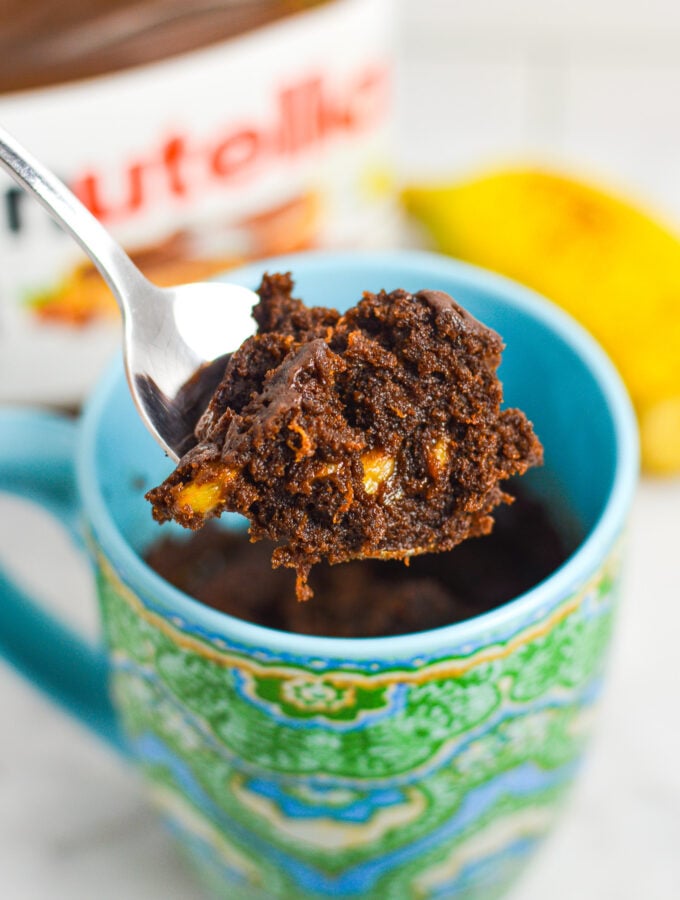 A small spoon taking a bite of Banana Nutella Mug Cake from a microwave safe mug, with the rich texture and small banana pieces showing in the chocolate cake.