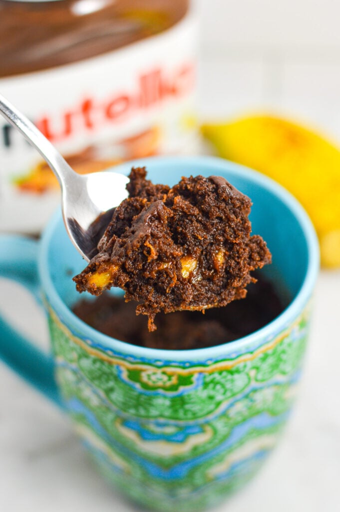 A small spoon taking a bite of Banana Nutella Mug Cake from a microwave safe mug, with the rich texture and small banana pieces showing in the chocolate cake.