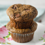 A stack of moist Double Chocolate Chip Muffins on a small plate, with semisweet chocolate chips sprinkled in the background.