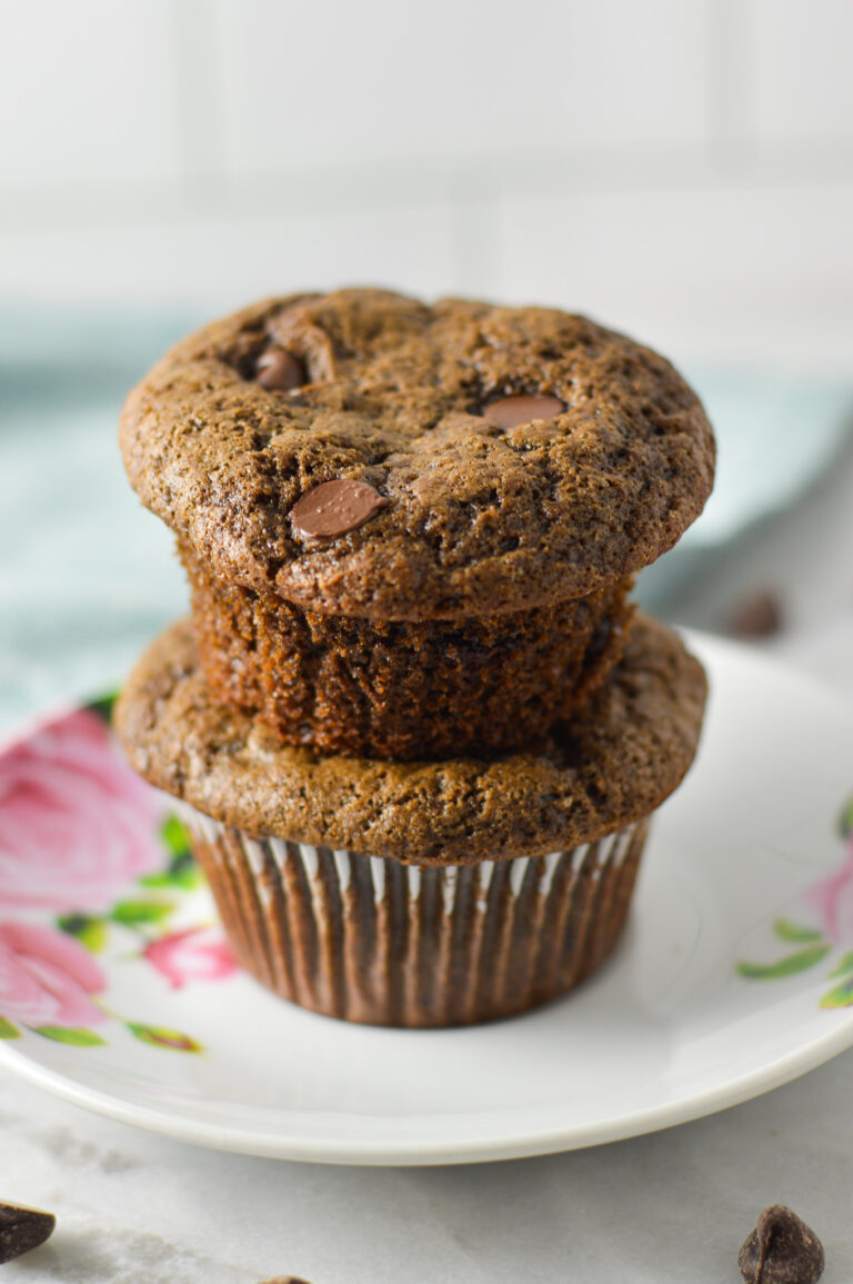 A stack of moist Double Chocolate Chip Muffins on a small plate, with semisweet chocolate chips sprinkled in the background.