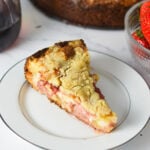 A slice of strawberry cream cheese coffee cake on a white plate, showing layers of soft butter cake, creamy cheesecake filling, vibrant strawberry sauce, and a golden crumb topping, with the whole cake just removed from a springform pan in the background