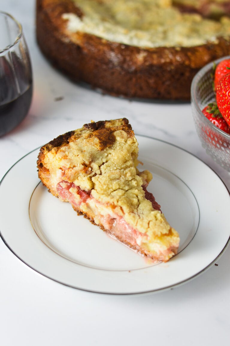 A slice of strawberry cream cheese coffee cake on a white plate, showing layers of soft butter cake, creamy cheesecake filling, vibrant strawberry sauce, and a golden crumb topping, with the whole cake just removed from a springform pan in the background