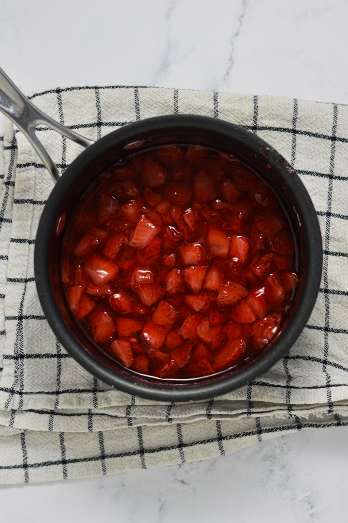 Strawberry sauce filling in a small saucepan on a white checkered dish towel.