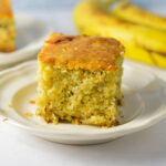 A slice of homemade Banana Vanilla Cake on a small plate, with some ripe bananas in the background.