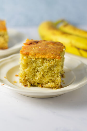 A slice of homemade Banana Vanilla Cake on a small plate, with some ripe bananas in the background.
