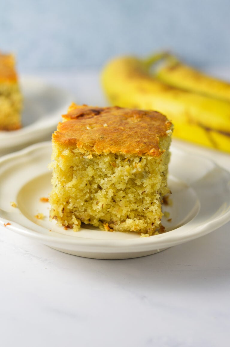 A slice of homemade Banana Vanilla Cake on a small plate, with some ripe bananas in the background.