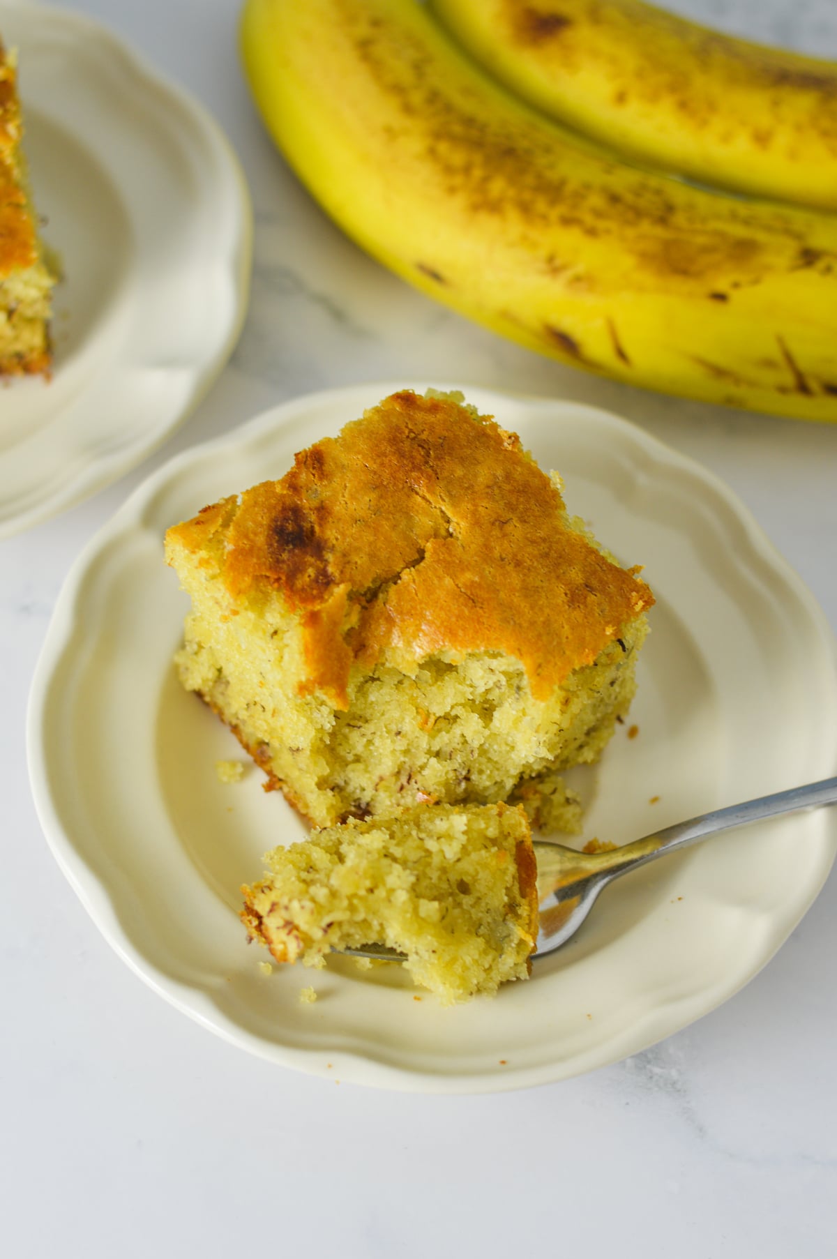 A square slice of Vanilla Banana Cake on a small plate with a fork taking a bite out of the homemade cake, with some brown spotted bananas in the background.