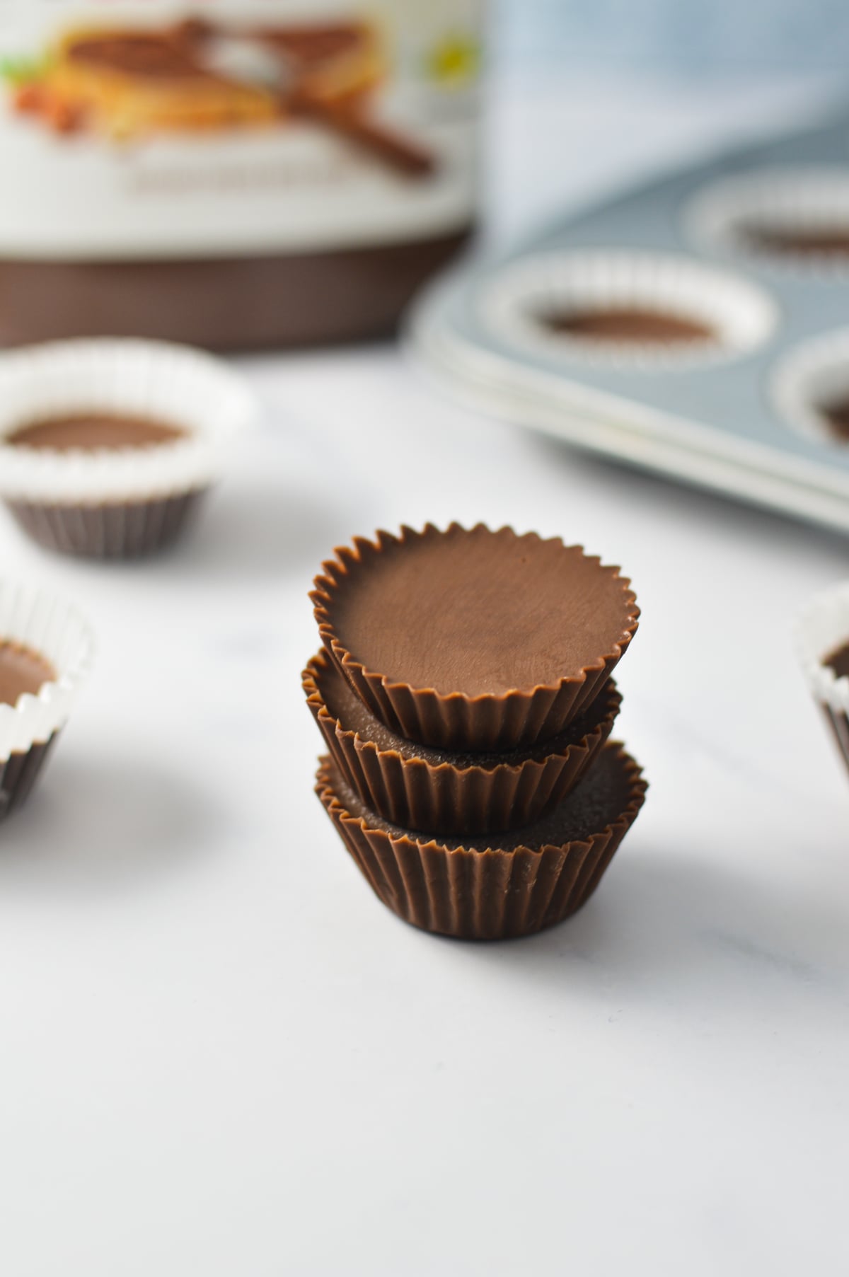 Three 2 Ingredient Nutella Fudge cups stacked on top of each other, with a jar of Nutella and a mini muffin pan filled with more Nutella cups in the background.