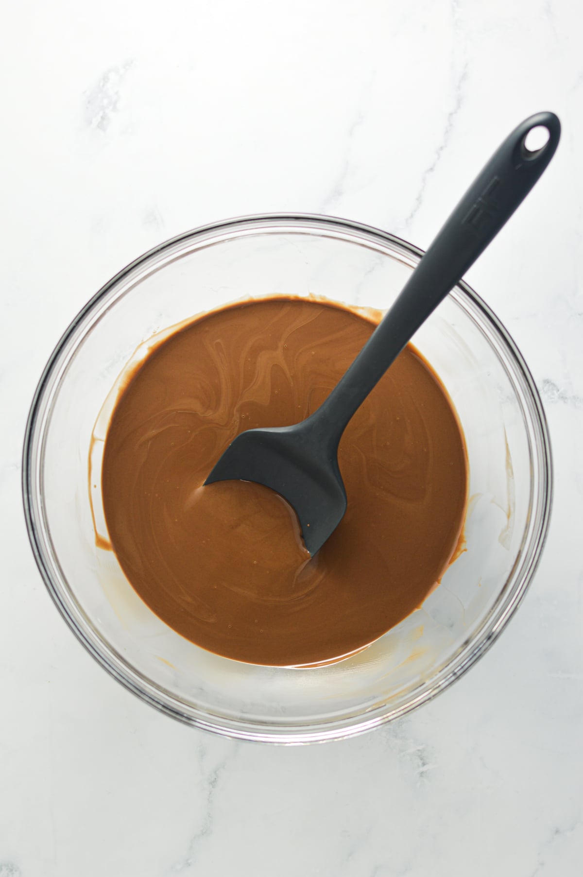 Melted Nutella and coconut oil mixed together in a large glass bowl with a silicone spatula inside.