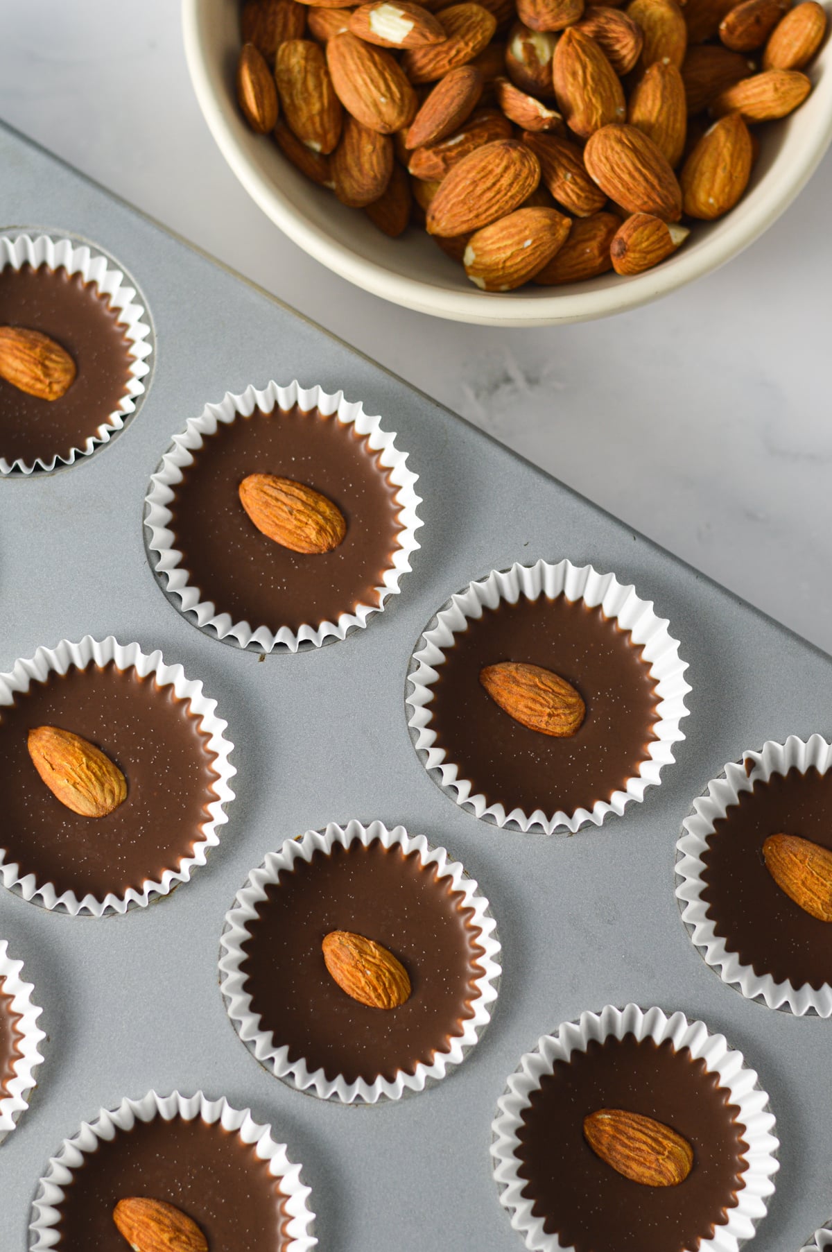A paper lined mini muffin pan filled with homemade Almond Mini Nutella Cups, with a small bowl of whole almonds beside it.