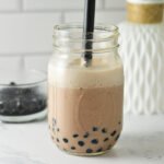 A mason jar filled with chocolate bubble tea and tapioca pearls, served with a black boba straw.