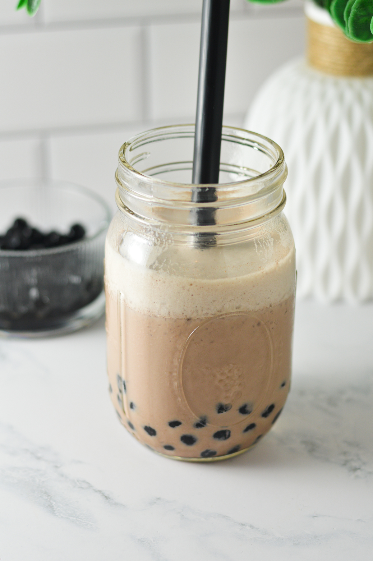 A glass mason jar containing chocolate bubble tea slushie over black boba pearls, served with a thick black straw against a light-colored background.