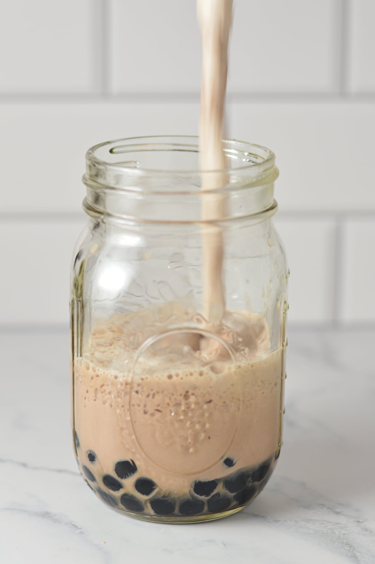 Creamy chocolate bubble tea being poured from a blender into a glass mason jar filled with black tapioca pearls at the bottom.