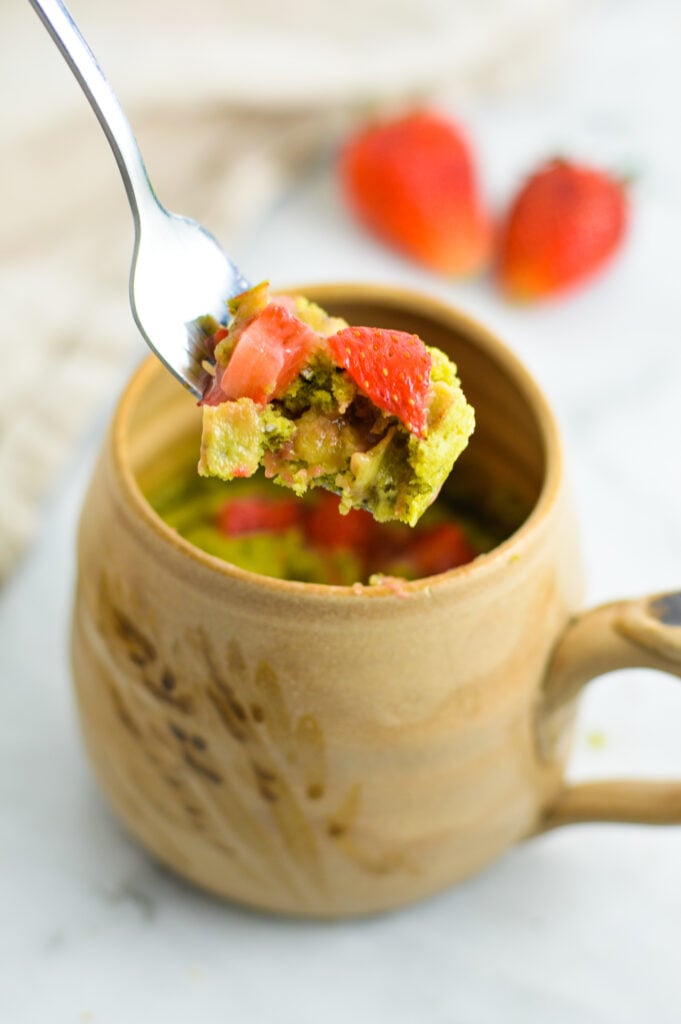 A fork full of Strawberry Matcha Mug Cake taken from a tan ceramic mug on a granite surface.