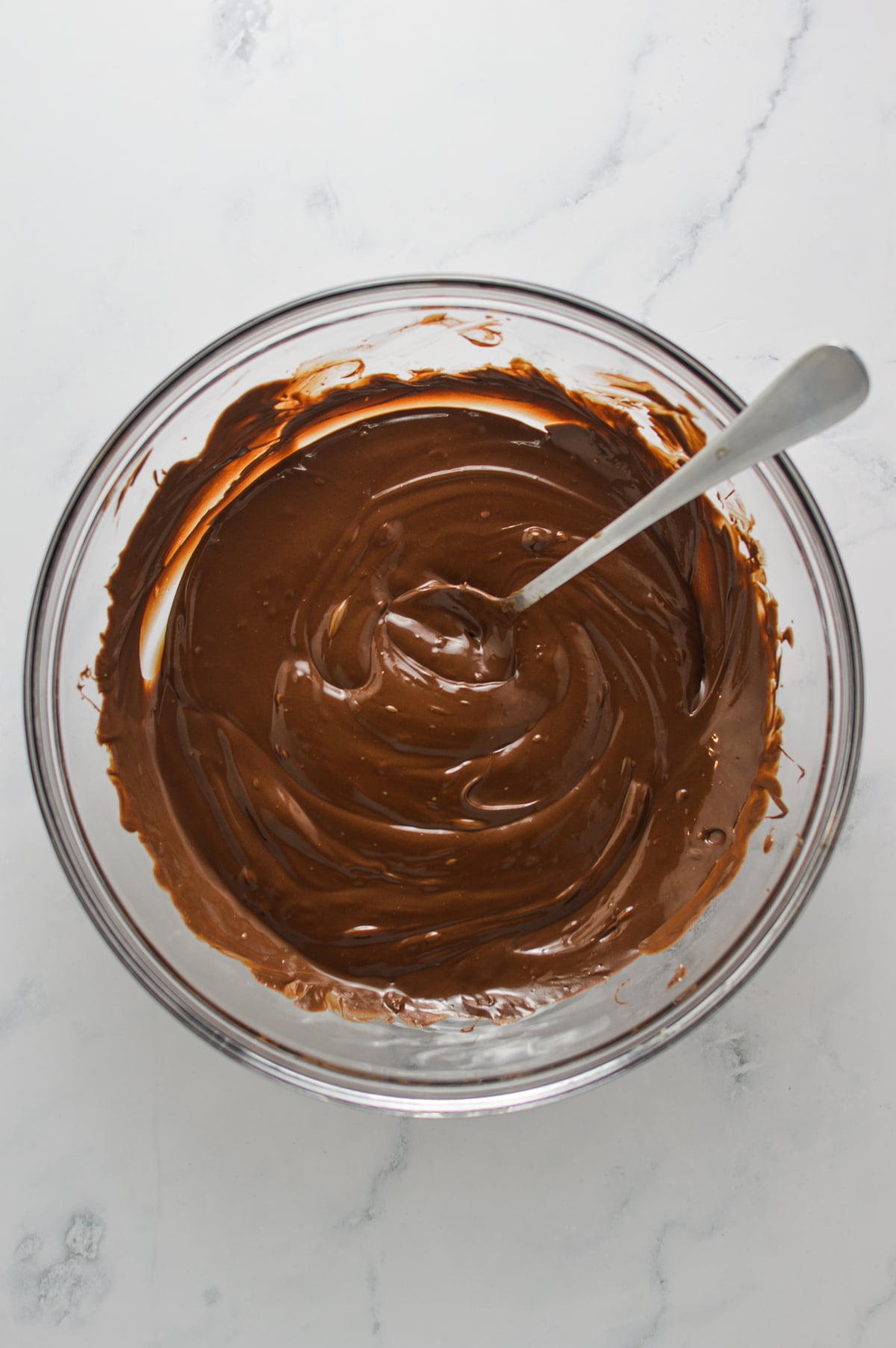 Chocolate chips and creamy chocolate hazelnut spread being mixed together with a spoon in a clear glass bowl.