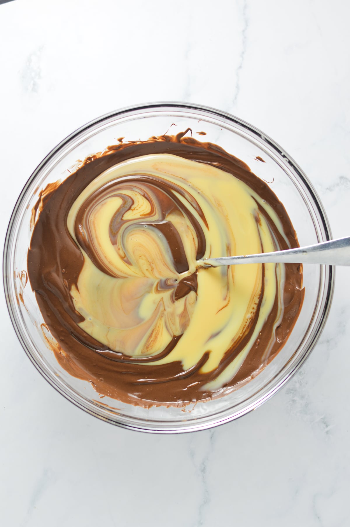 Sweetened condensed milk being poured into a chocolate and Nutella mixture in a glass bowl, creating a light and dark brown swirl.