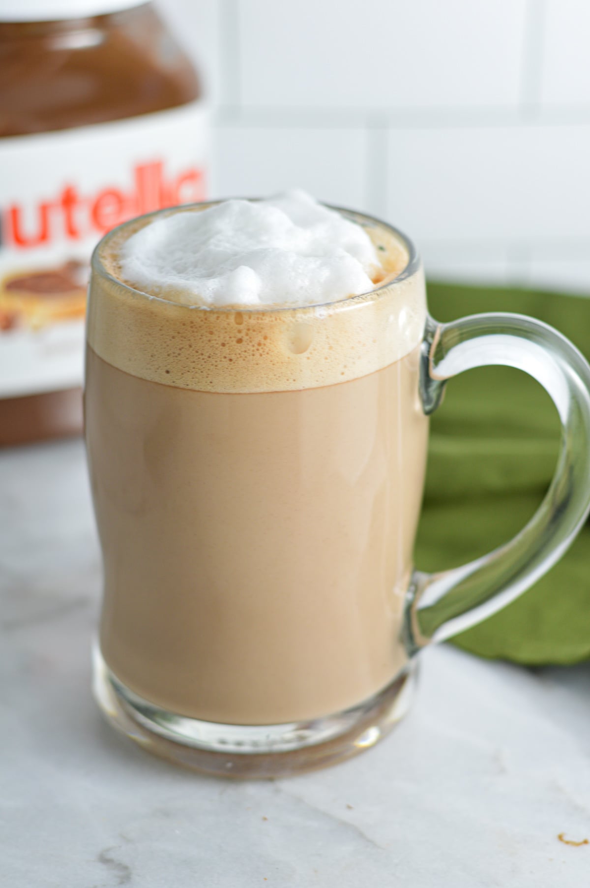 Nutella Latte in a glass mug with foamed milk on top and a jar of Nutella behind the rich drink.