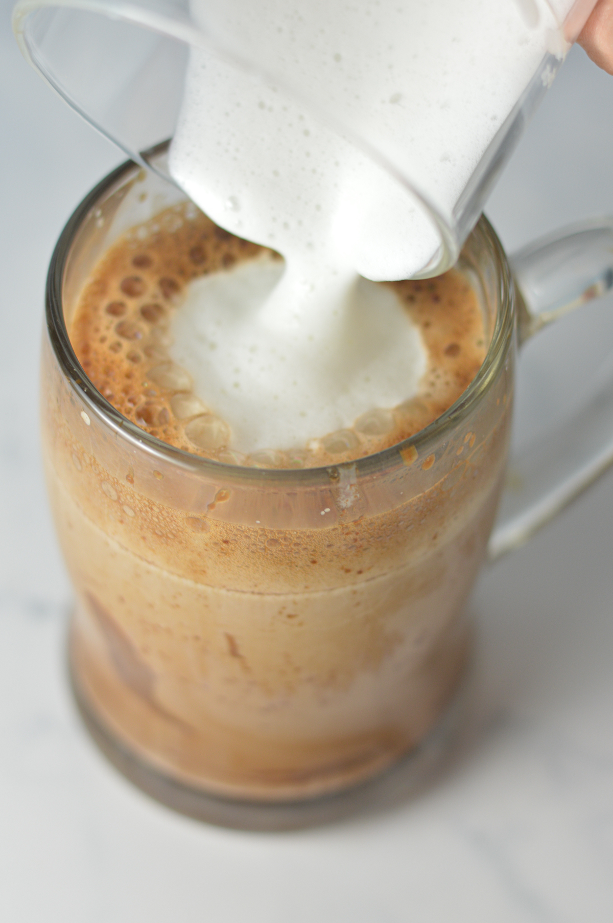 A hand pouring some frothed milk overtop a clear mug full of Nutella Latte.