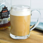 Maple Latte in a glass mug with a jar of maple syrup in the background.