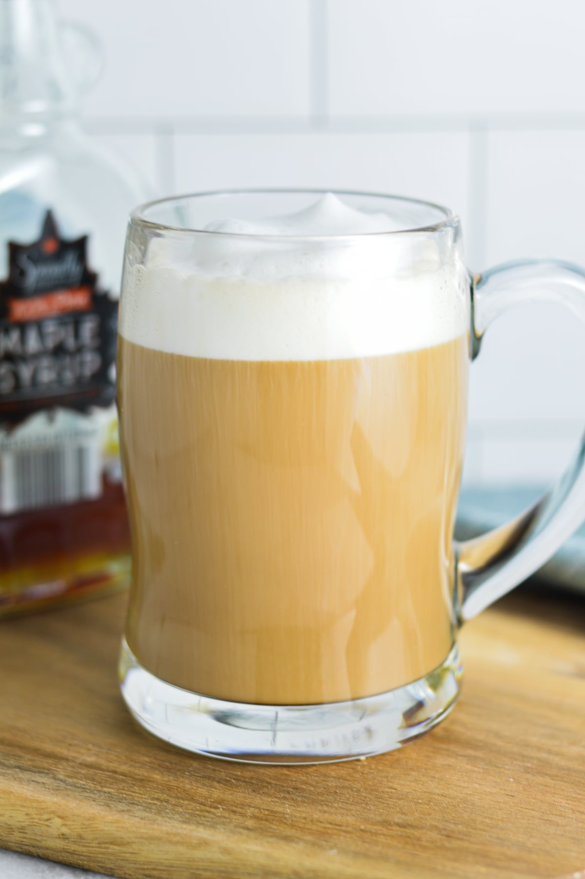 A coffee mug filled with a homemade Maple Latte served on a wooden surface, with a jug of pure maple syrup in the background.