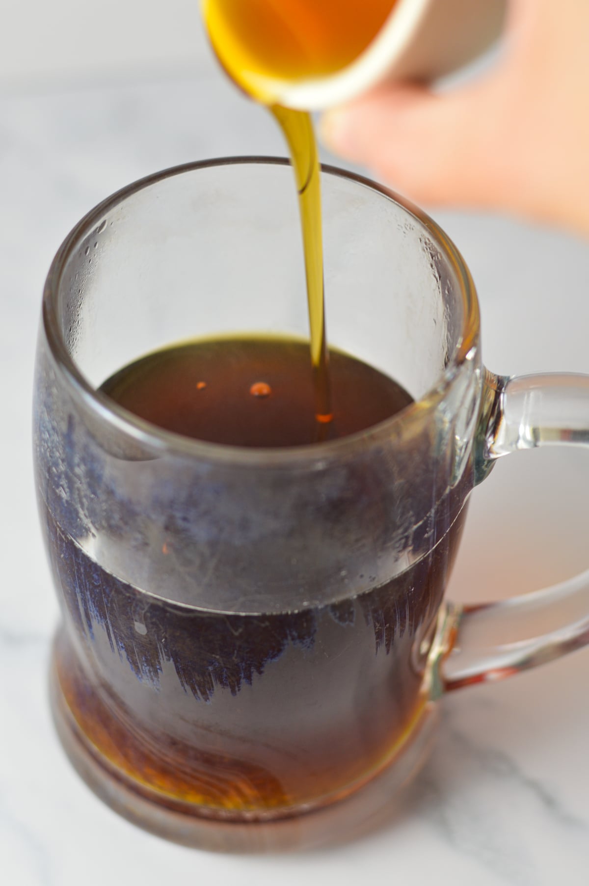 Some pure maple syrup being poured into a glass mug with strong brewed coffee in it.