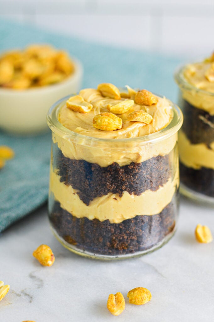A closeup of two Peanut Butter Chocolate Cake Cheesecake Cups. Each cup is layered with moist chocolate cake and creamy peanut butter cheesecake, and topped with dry roasted peanuts.